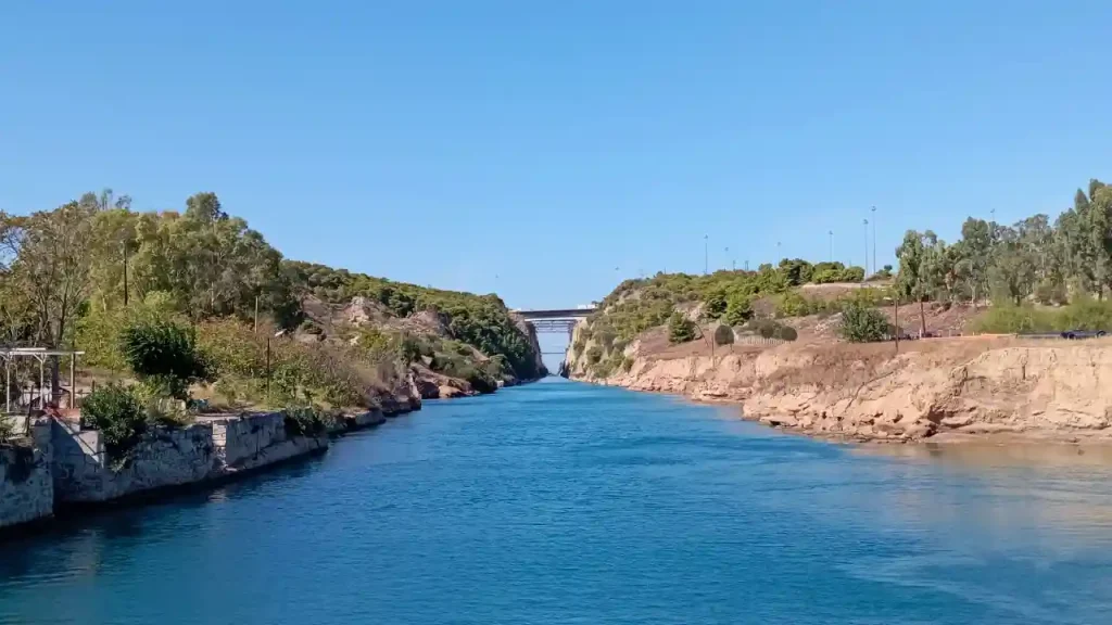 A panoramic view of the Corinth Canal, a man-made waterway in Greece. The canal cuts through the Isthmus of Corinth, connecting the Gulf of Corinth to the Saronic Gulf. The canal is flanked by rocky cliffs and lush greenery, with a bridge spanning across it.