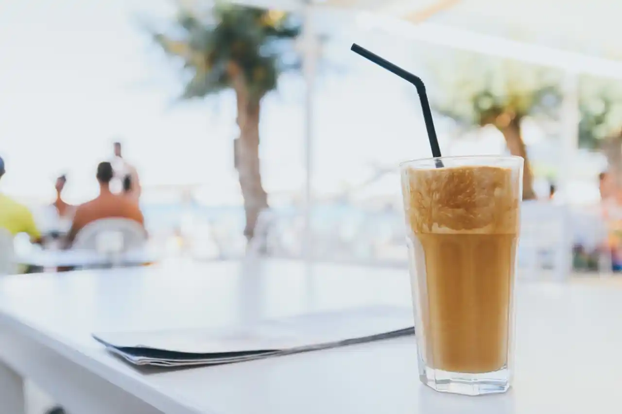 A tall glass of frothy Frappe coffee with a straw, sitting on a white table. A newspaper is next to the glass, and there is a blurred background of people sitting at tables on a patio.