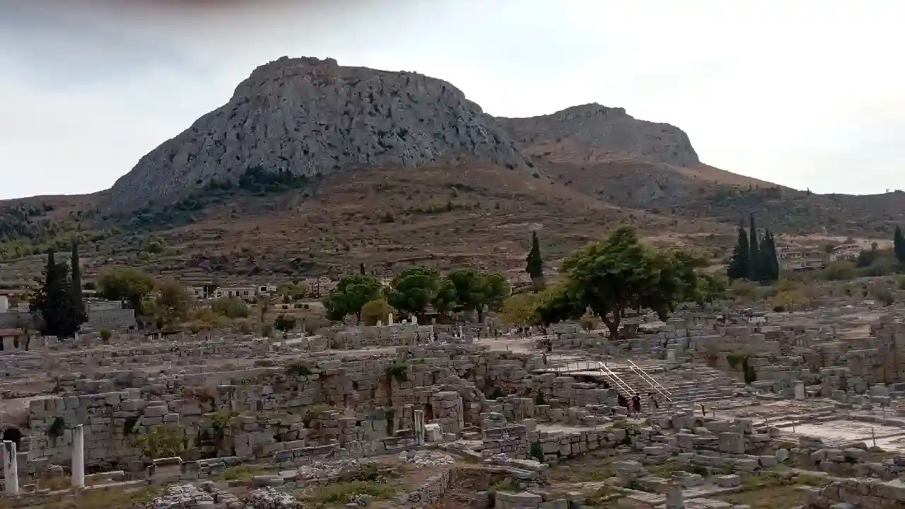 A panoramic view of the ancient city of Corinth, Greece. The ruins of the city are spread out below, with the towering Acrocorinth hill in the background.