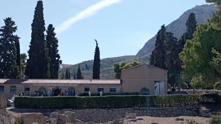 A photo of the Archaeological Museum of Ancient Corinth, Greece. The museum building is a low-slung structure with a terracotta roof, surrounded by cypress trees and ancient ruins. The Acrocorinth mountain is visible in the background.