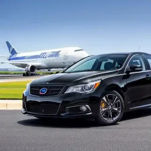 Athens Airport Transfer. A black Subaru Legacy sedan parked in front of a passenger airplane on a runway. The airplane appears to be a private jet.