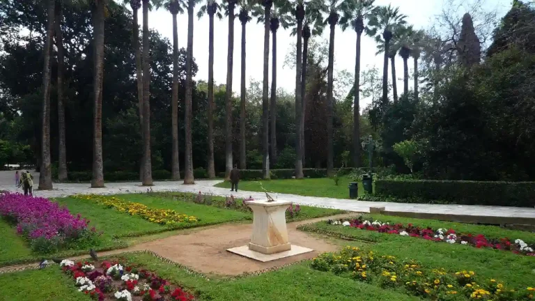 A scenic view of the National Gardens in Athens, Greece. Tall palm trees line the paths, and colorful flower beds dot the landscape. A sundial stands in the center, surrounded by lush greenery.