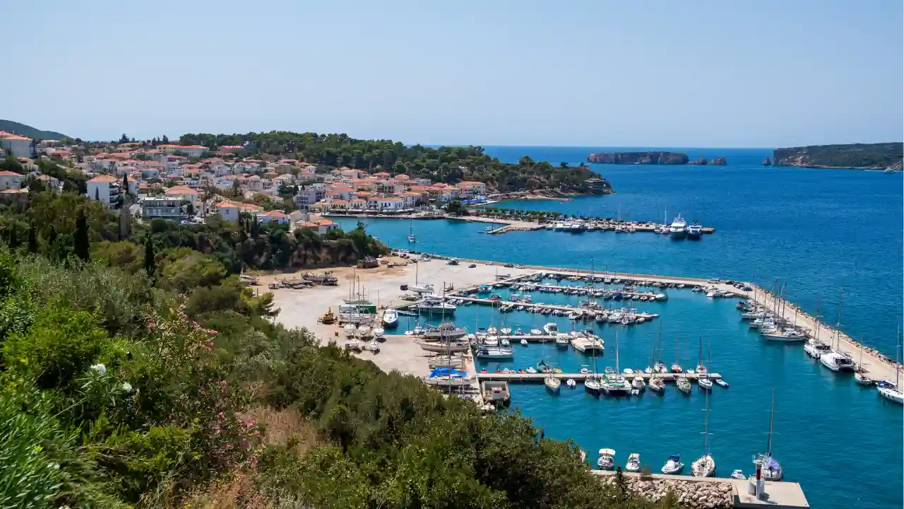 A photo of a marina with sailboats and yachts docked in the water. The marina is surrounded by hills and mountains. In the background is a small town.