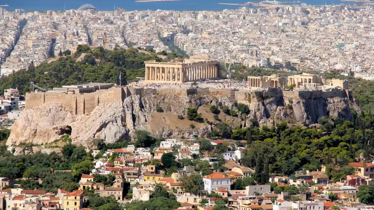 A panoramic view of the Acropolis of Athens, Greece, with the Parthenon prominently displayed. The city of Athens stretches out below, with the Aegean Sea visible in the distance.