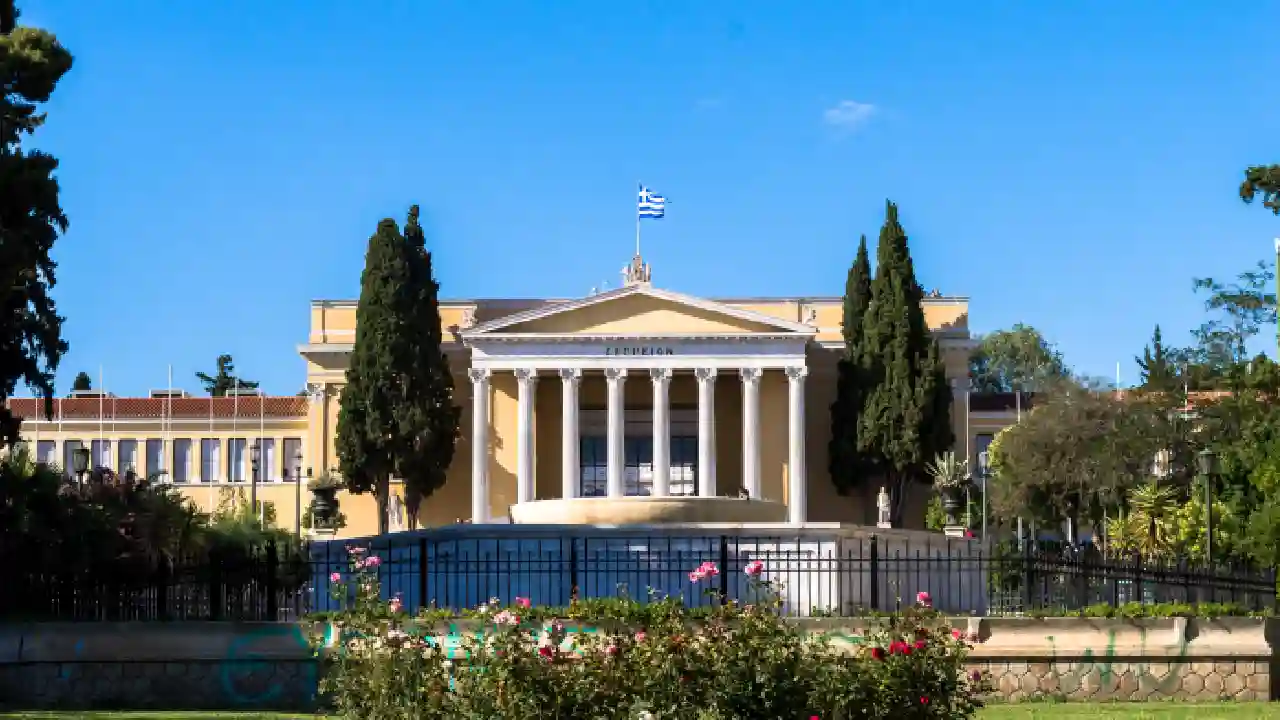 Close-up of the Zappeion's main entrance. The entrance is made of white marble and features a large Corinthian portico. The Greek flag is flying above the entrance.