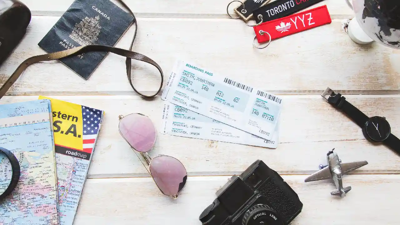 A close-up photo of a wooden table with travel essentials laid out on top. There is a passport, boarding passes, sunglasses, a camera, and a folded map.