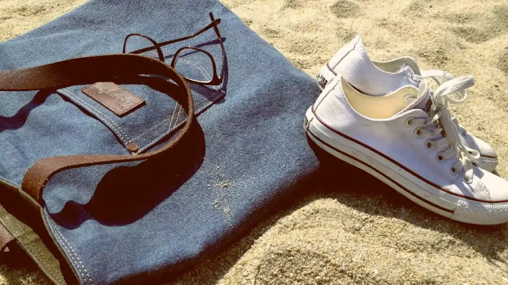 A pair of white sneakers next to a denim tote bag and sunglasses on a beach.