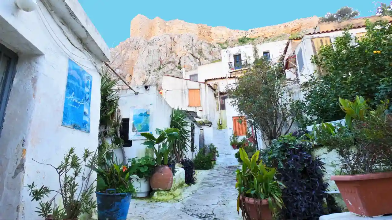 A small alleyway with brightly colored houses on either side. There are potted plants overflowing with flowers lining the alleyway, and a flight of stairs leads upwards in the distance. In the background, a lush green mountain can be seen. This is Anafiotika, a historic neighborhood perched on a hill in Athens, Greece.