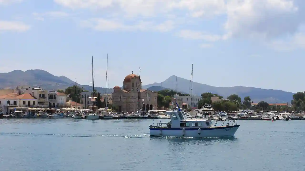 Aegina. A small wooden boat floating on calm blue water in a harbor. In the background are colorful buildings, a white church, and green mountains