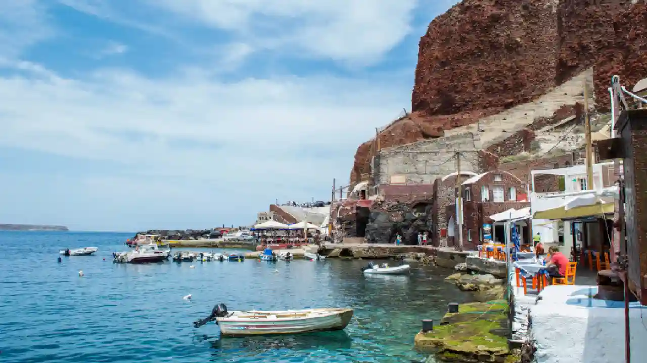 A scenic harbor with traditional fishing boats resting at the dock. The whitewashed houses with colorful accents in the background add to the charm of the Greek landscape.