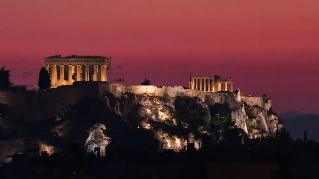 A view of the Acropolis , illuminated by floodlights. The Parthenon is the most prominent building on the hilltop. In the foreground are some dark trees and shrubs.