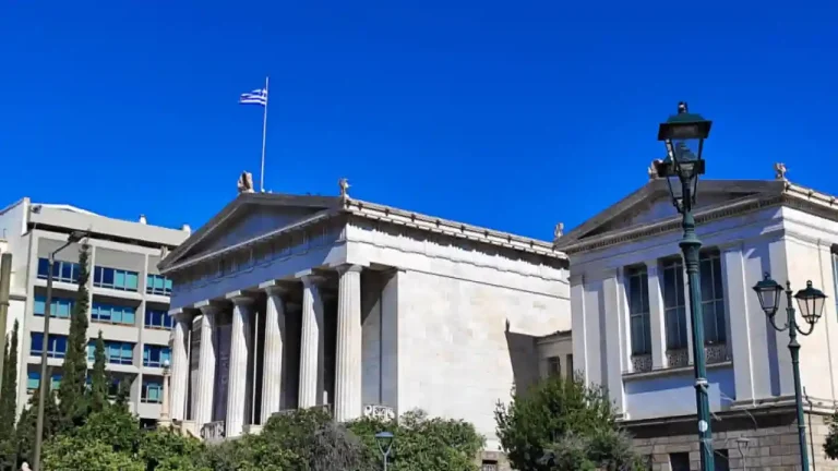 The Vallianeio Megaron, the National Library of Greece, located on Panepistimiou Avenue in Athens. The building is neoclassical with a portico of six columns and a pediment adorned with sculptures. There are modern buildings in the background and a lamppost on the right side.