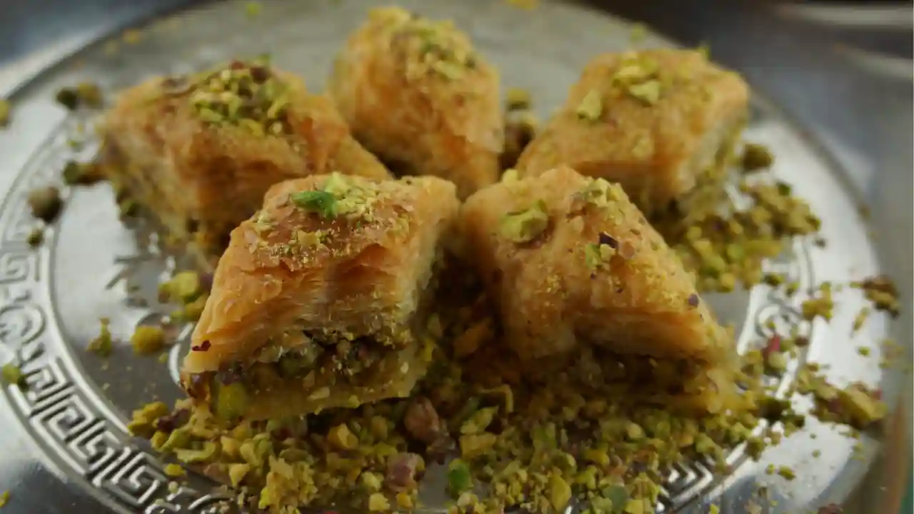 A close-up of a plate of Greek baklava. The baklava is made with filo dough, chopped nuts, and sweetened with syrup. It is topped with pistachios.