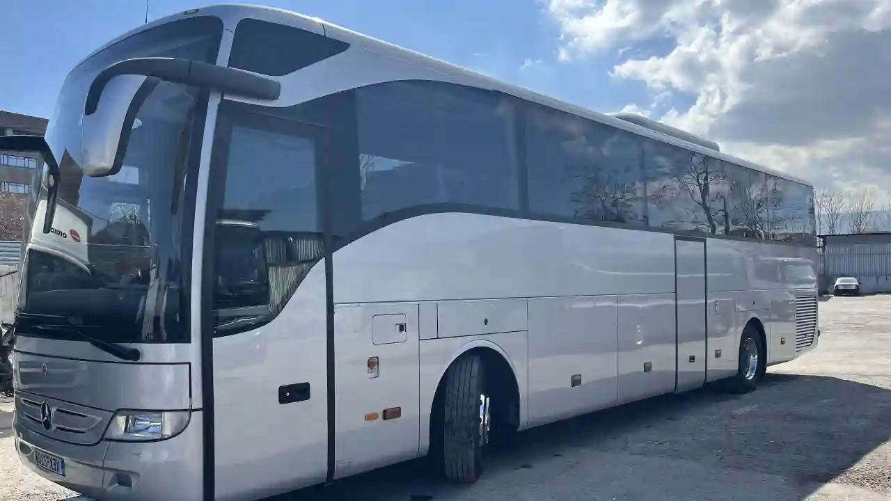 A white Mercedes-Benz Tourismo coach bus parked in a lot with a clear blue sky and some clouds.