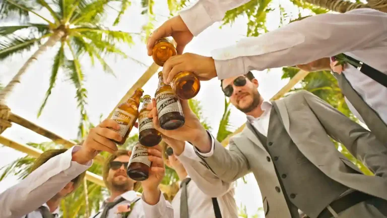 A group of men dressed in suits celebrating with a toast of beer bottles under palm trees, enjoying a bachelor party.