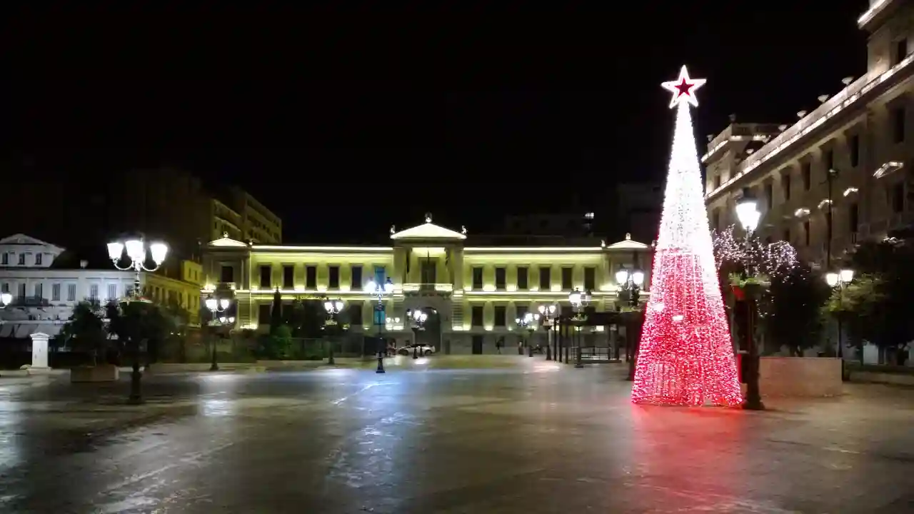 A brightly lit Christmas tree stands in a public square in Athens, Greece. The National Bank of Greece building is illuminated in the background, and the square is decorated with festive lights