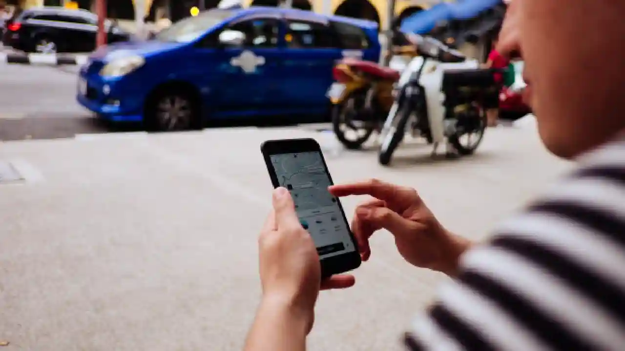 A person using Uber application in a smartphone while standing on a sidewalk next to a parked motorcycle and blue car.