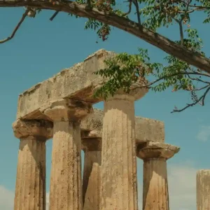 Ruins of the Temple of Apollo in Corinth, Greece.. The temple is made of white marble and has six columns on each side. The sky is blue and there are a few clouds in the background. The photo is taken from the ground level, looking up at the temple.
