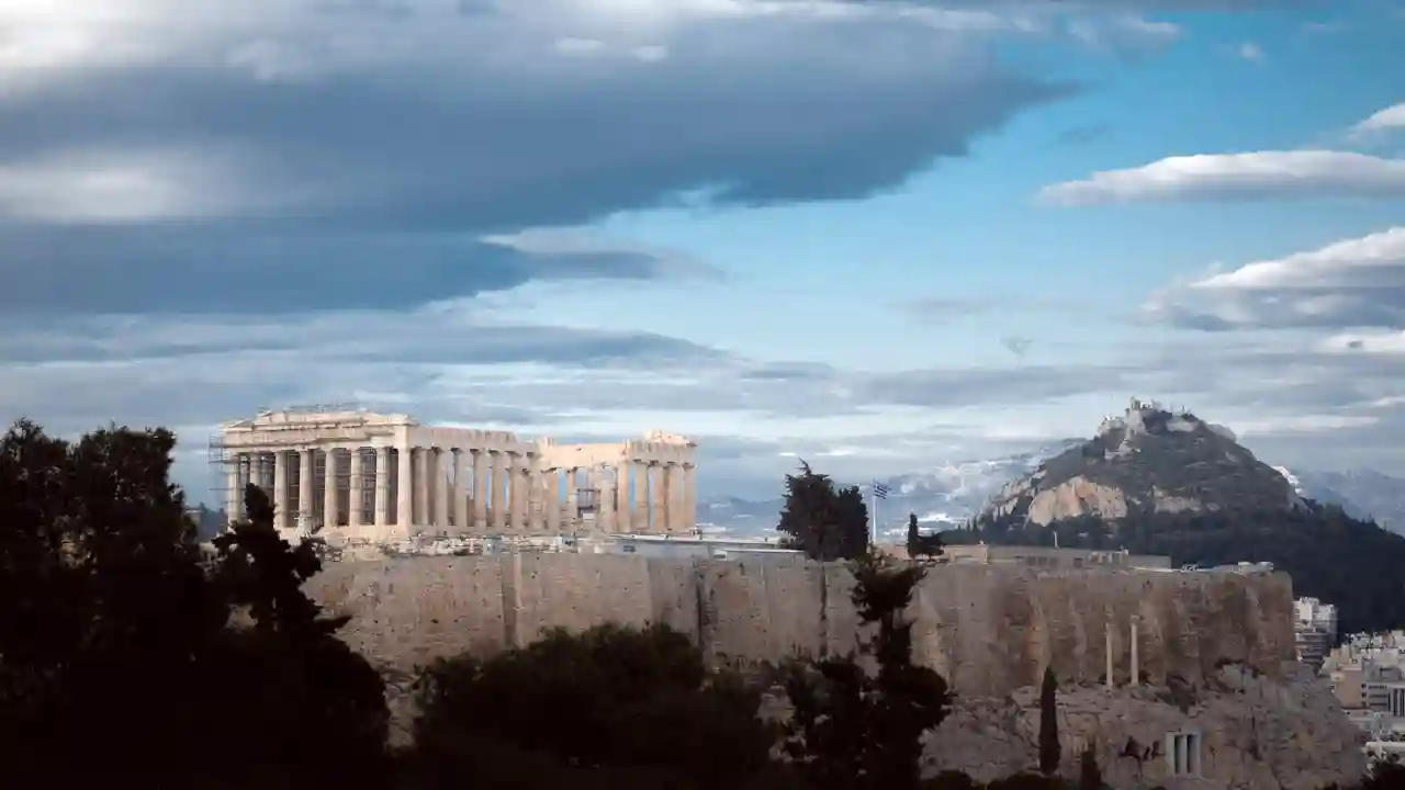 A panoramic view of the Acropolis of Athens, Greece in December. The Parthenon, a large white marble temple dedicated to the goddess Athena, is the centerpiece of the image. The temple is surrounded by other ancient ruins, and the city of Athens can be seen in the background. The sky is cloudy, and there is a mountain range in the distance.