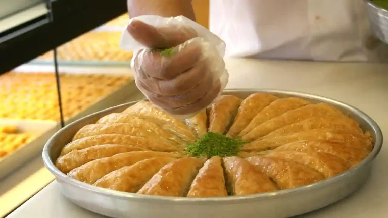 A gloved hand sprinkles chopped pistachios on top of a freshly baked Greek baklava. The baklava is arranged in a circular pattern in a metal tray. The phyllo dough is golden brown and flaky, and the pistachios are a bright green. The baklava is a delicious and popular Greek dessert.