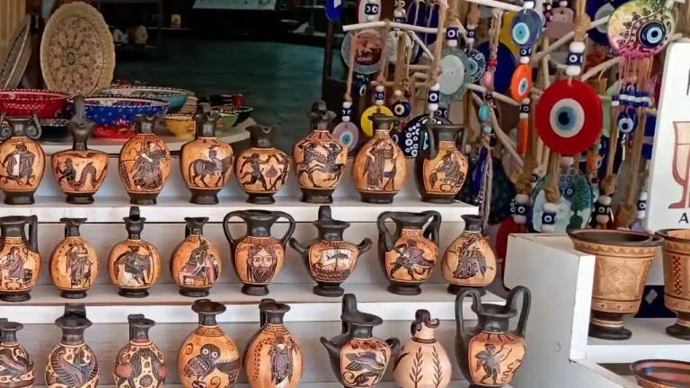 A display of traditional Greek souvenirs, including pottery vases with intricate black-figure designs depicting mythological scenes, evil eye charms, and other handcrafted items.