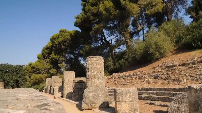 Ruins of the Temple of Zeus at Olympia, Greece. The photo shows rows of weathered columns and stone foundations amidst lush greenery and tall trees. The remains of the ancient temple stand against a clear blue sky.