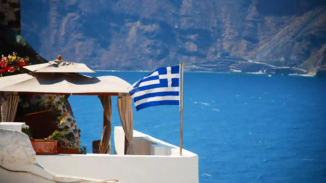 A Greek flag waving in the wind on a white balcony overlooking the Aegean Sea. The sky is blue and there are no clouds. The sea is calm and there is a small island in the distance.