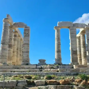 Ruins of the Temple of Poseidon at Cape Sounion, Greece. The temple stands on a cliff overlooking the Aegean Sea. The columns are weathered and the sky is a clear blue.