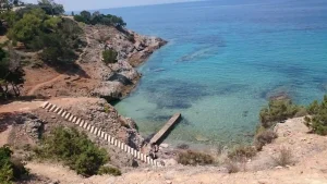 A secluded cove with crystal-clear turquoise water and rocky cliffs. A staircase leads down to a small wooden pier. The sky is a clear blue.