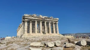 The Parthenon, a historic temple on the Acropolis in Athens, Greece, undergoing restoration work. Scaffolding surrounds the structure, with clear blue skies above.