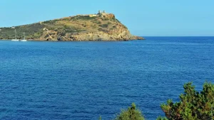 A panoramic view of the Temple of Poseidon at Cape Sounion, Greece. The temple stands on a rocky promontory overlooking the Aegean Sea. The water is a deep blue and there are sailboats visible in the distance.