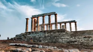 The Temple of Poseidon at Cape Sounion, Greece. The temple is a historic ruin with standing columns overlooking the Aegean Sea. The sky is a dramatic blue with clouds.