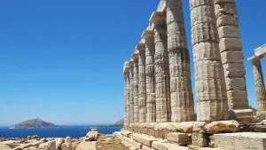 A close-up view of the columns of the Temple of Poseidon at Cape Sounion, Greece. The columns are weathered and stand tall against a clear blue sky. The Aegean Sea is visible in the background.