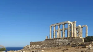 A panoramic view of the Temple of Poseidon at Cape Sounion, Greece. The temple is a historic ruin with standing columns overlooking the Aegean Sea. The sky is a clear blue.