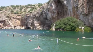 Vouliagmeni lake surrounded by cliffs, with people swimming and sunbathing. The water is a vibrant blue-green, and there are buoys marking a swimming area.