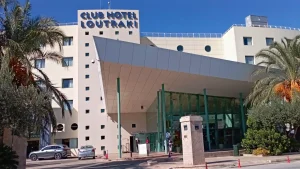 Club Hotel Loutraki, a modern hotel in Loutraki, Greece, featuring a casino. The hotel has a white facade with blue accents and palm trees in front