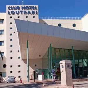 Club Hotel Loutraki, a modern hotel in Loutraki, Greece, featuring a casino. The hotel has a white facade with blue accents and palm trees in front