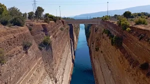 A bridge spanning the Corinth Canal, a man-made waterway in Greece. The canal cuts through the Isthmus of Corinth, connecting the Gulf of Corinth to the Saronic Gulf. The canal is flanked by rocky cliffs and lush greenery.