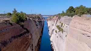 A view of the Corinth Canal from the west side of the bridge. The canal cuts through the Isthmus of Corinth, connecting the Gulf of Corinth to the Saronic Gulf. The canal is flanked by rocky cliffs and lush greenery.