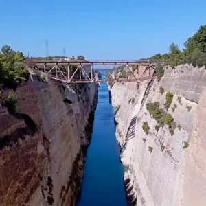 A view of the Corinth Canal from the west side of the bridge. The canal cuts through the Isthmus of Corinth, connecting the Gulf of Corinth to the Saronic Gulf. The canal is flanked by rocky cliffs and lush greenery.