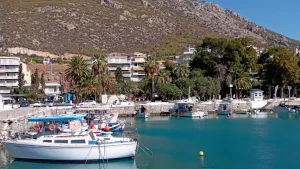 A picturesque harbor scene in Loutraki City, Greece. Several fishing boats are docked in the turquoise waters, surrounded by palm trees and white buildings. A mountain range rises in the background.