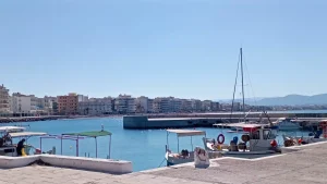 A scenic harbor in Loutraki, Greece. Several fishing boats are docked at the pier, with a sailboat in the background. The harbor is surrounded by buildings and a mountain range.