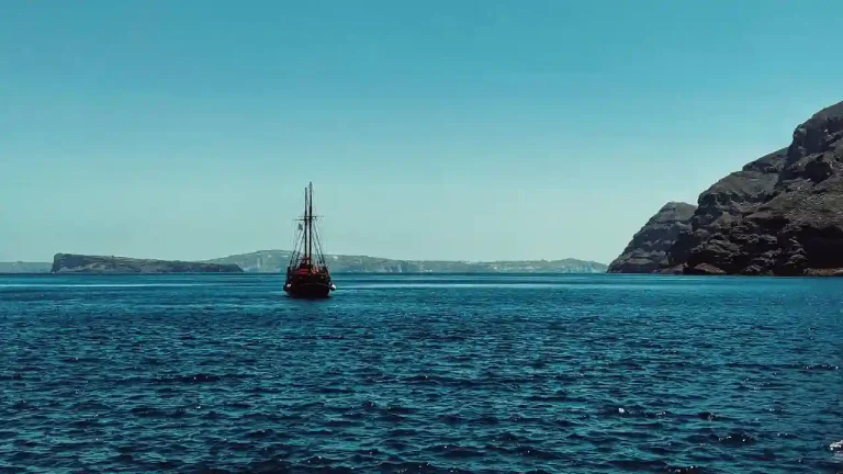 A traditional Greek sailboat, painted in a deep red hue, sails across the crystal-clear waters of the Aegean Sea. The boat is silhouetted against a bright blue sky, with the iconic cliffs of Santorini in the background.