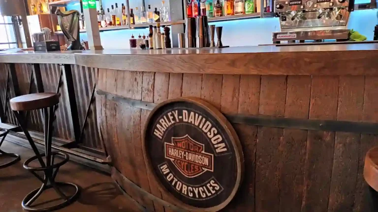 A stylish bar counter with a Harley-Davidson logo prominently displayed, showcasing the brand's connection to the motorcycle culture. The bar is well-stocked with various spirits and coffee machines, creating an inviting atmosphere for motorcycle enthusiasts and cocktail lovers alike.