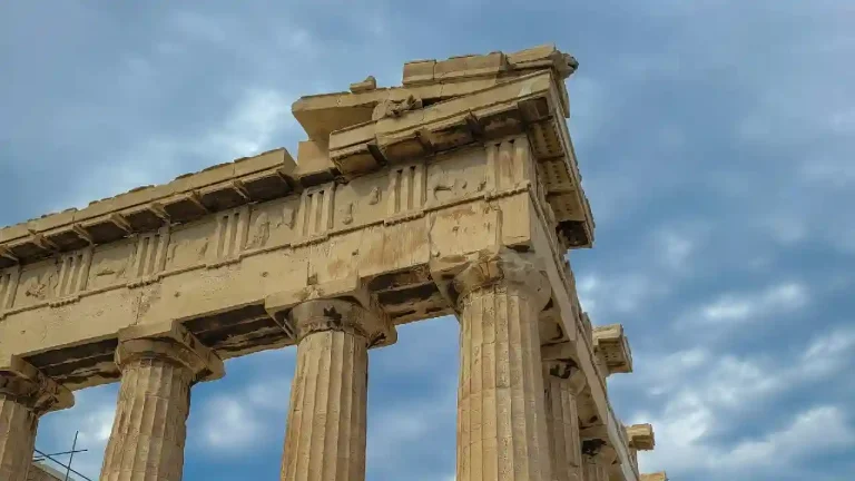 A close-up view of the Parthenon, an ancient Greek temple on the Acropolis in Athens. The image showcases the intricate details of the temple's columns, frieze, and pediment against a dramatic cloudy sky.