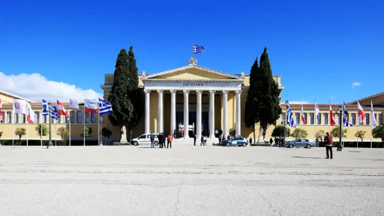 Zappeion Hall, a grand neoclassical building in Athens, Greece, stands tall under a clear blue sky. The building is adorned with flags from various countries, creating a festive atmosphere.