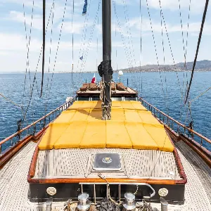 A spacious sun deck on a traditional Greek sailing yacht, with comfortable yellow cushions and a panoramic view of the Aegean Sea. The yacht is sailing towards a distant island, promising an unforgettable island-hopping adventure.