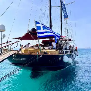 A large wooden sailing yacht named "Angelique" docked at a pier in Greece. The Greek flag is flying proudly from the mast, and people are gathered on the deck enjoying the beautiful scenery.