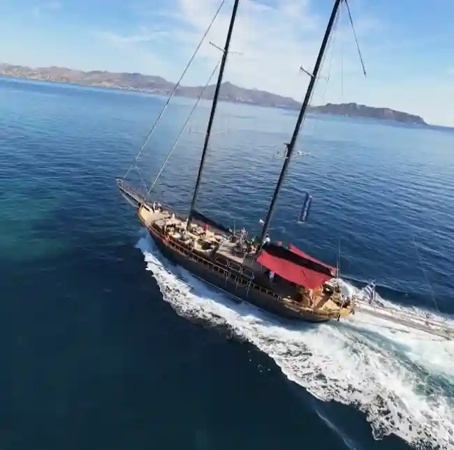 A large wooden sailing yacht with two masts, sailing through the crystal-clear blue waters of the Aegean Sea. The yacht is cutting through the water, leaving a trail of foam in its wake.