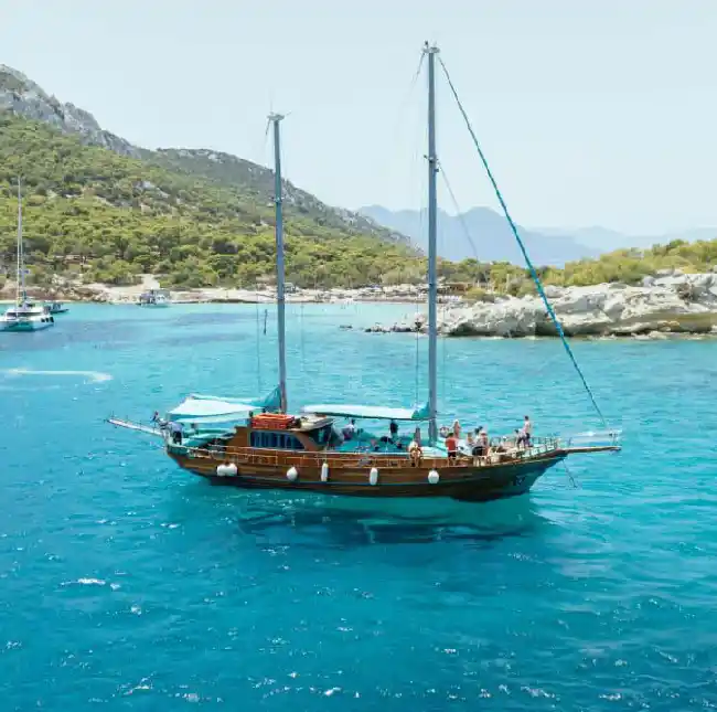 A large wooden sailing boat with two masts, anchored in a crystal-clear turquoise bay. Lush green hills surround the bay, and other boats are visible in the distance.